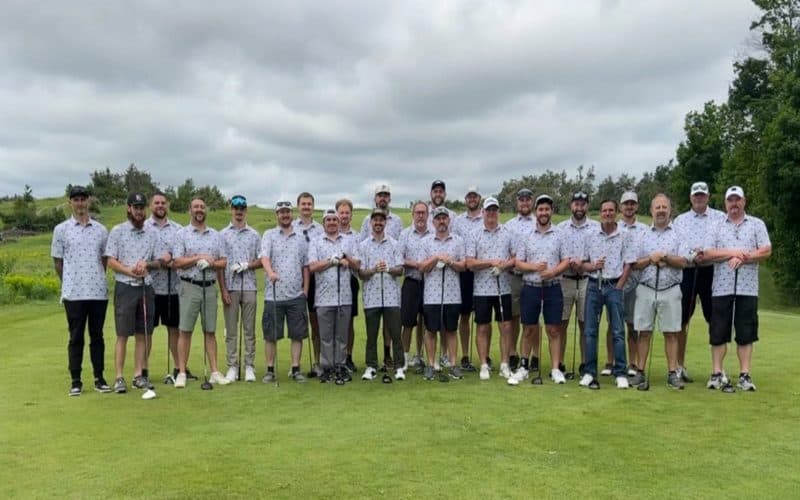 A team of about twenty golfers in matching white polos with a small all-over pattern, posing together on a golf course.