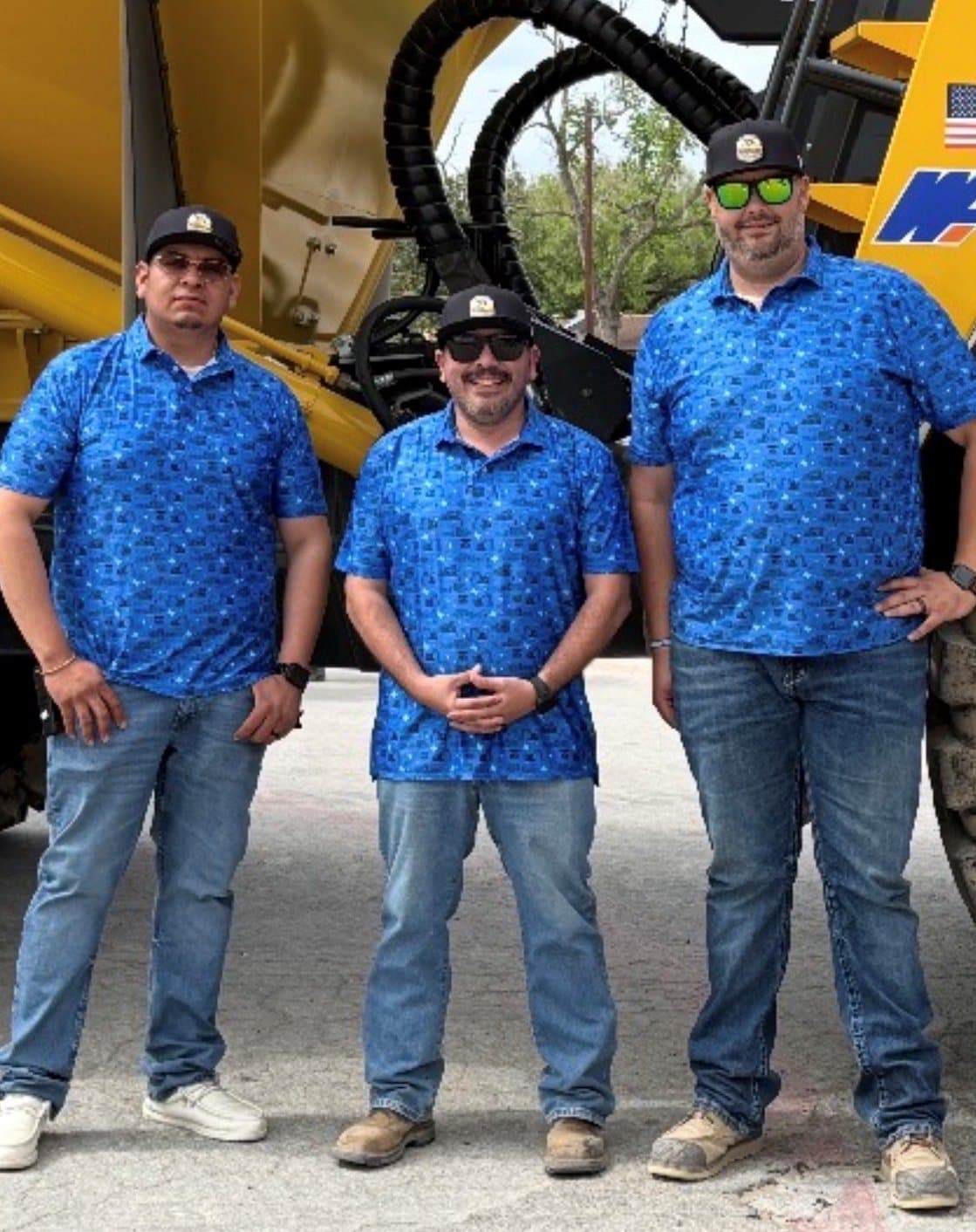 Three equipment-yard crew members in matching blue custom polos in front of heavy equipment