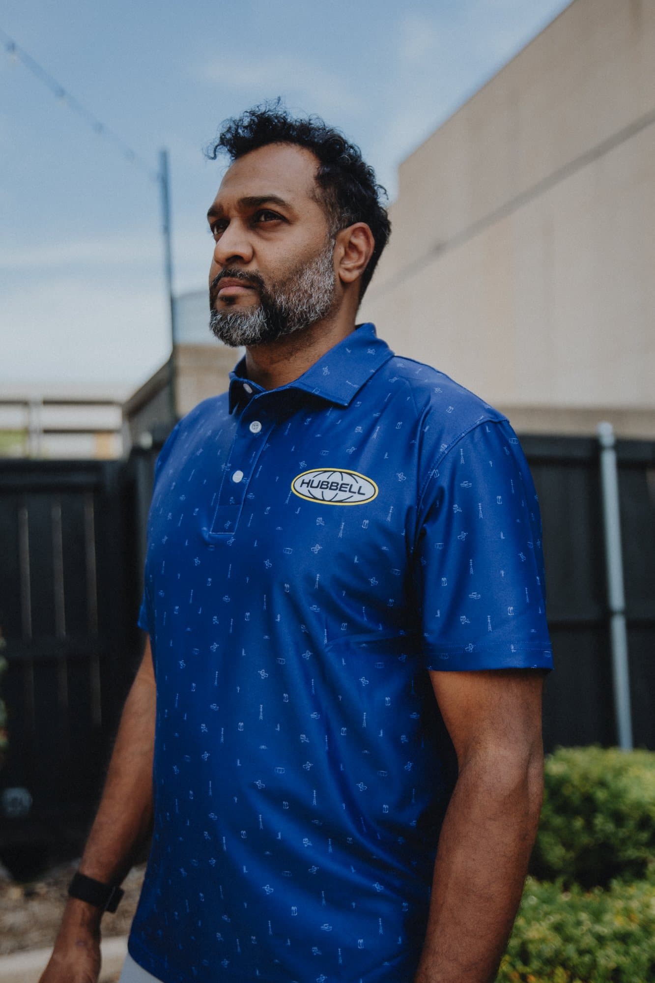Man in a royal blue polo with an oval chest badge standing outdoors
