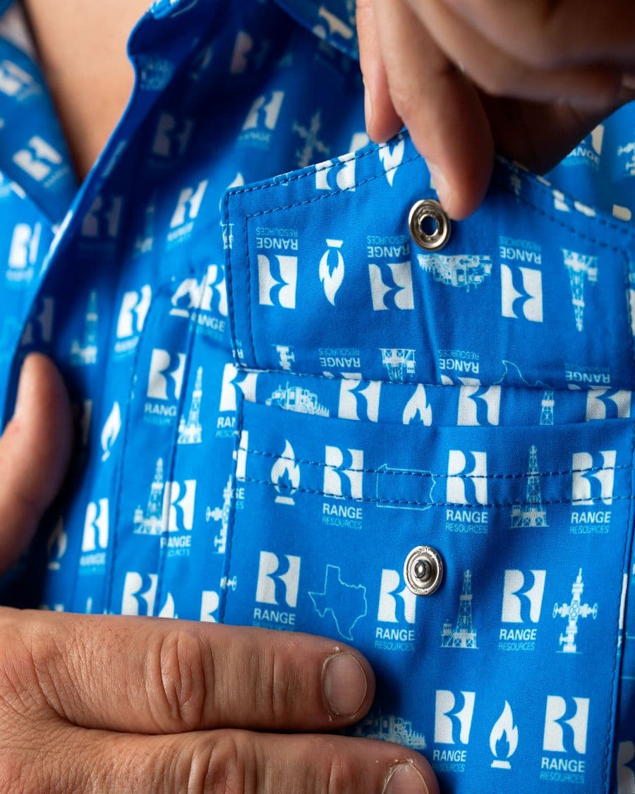 A hand lifting the flap of a chest pocket on a royal blue button-up to show the snap on the flap and the snap on the pocket.