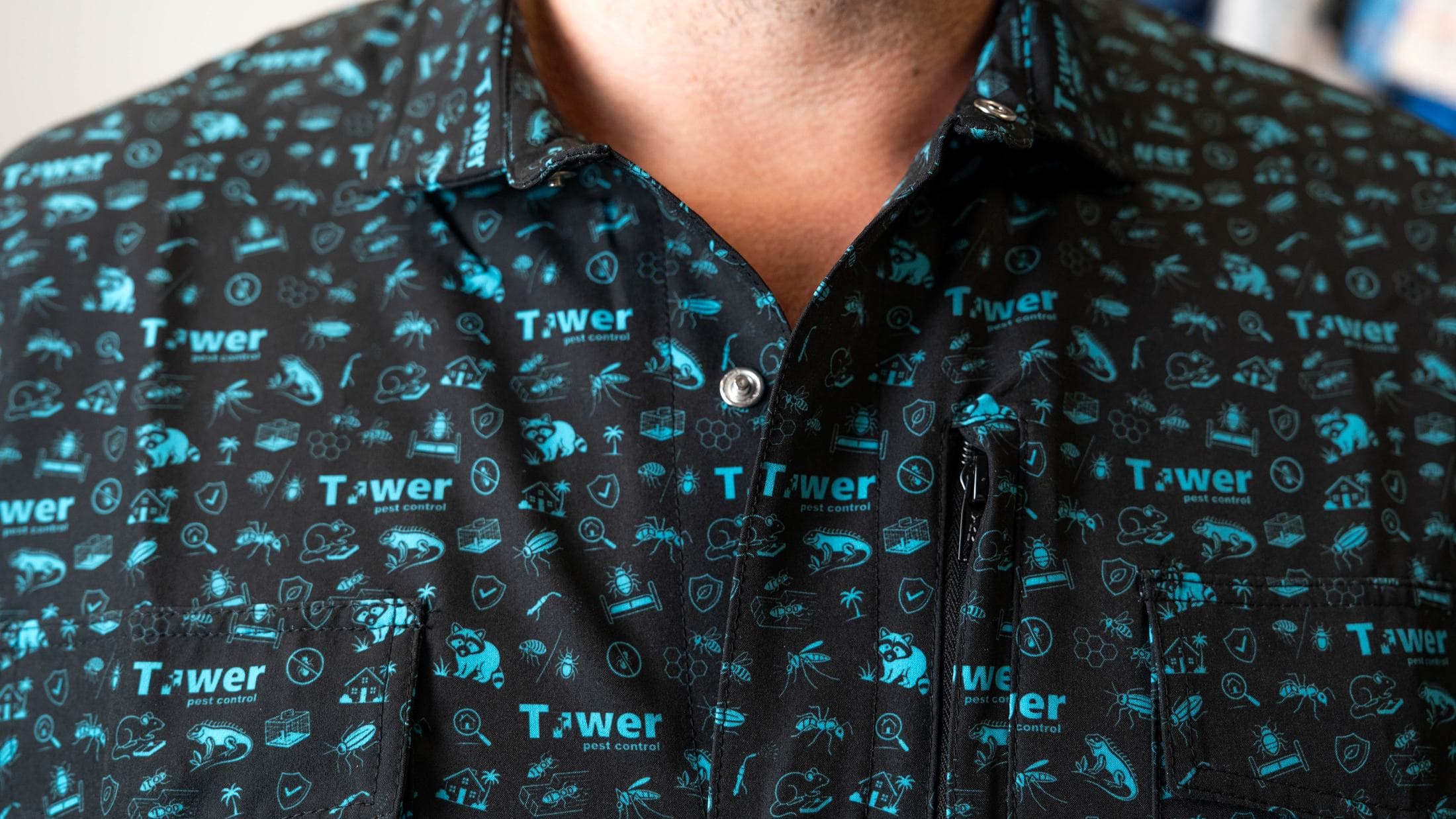Close-up of a black button-up with a teal all-over print, showing the snap at the collar, a snap on the placket, a zip pocket and flap pockets.