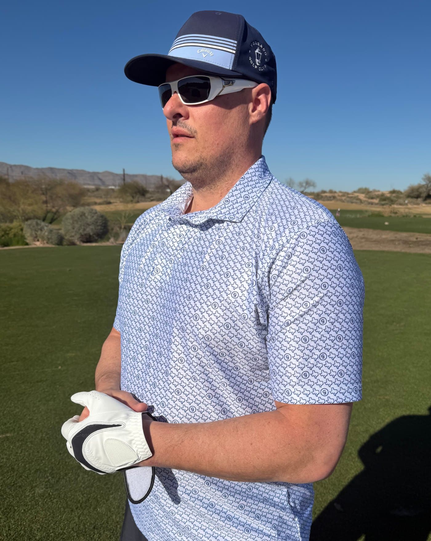 A golfer in sunglasses and a cap wearing a white all-over print polo on a sunny course