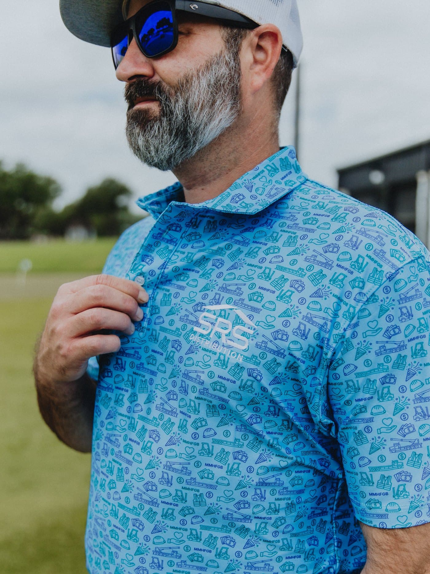 A bearded man in sunglasses and a trucker cap adjusting the collar of a light blue all-over print polo