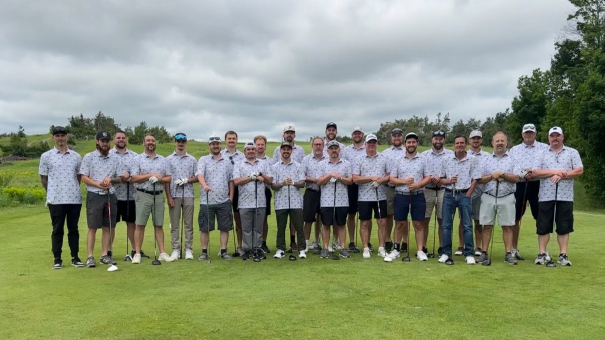 A team of about twenty golfers in matching white polos with a small all-over pattern, posing together on a golf course before a tournament.